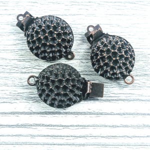 May include: Three round metal jewelry clasps featuring a dark, oxidized copper finish and a textured, honeycomb-like surface pattern. Each clasp includes a small circular loop at one end and a flat, hinged mechanism for securing items.
