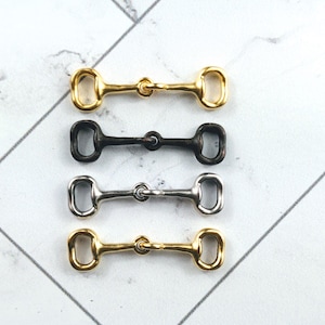 May include: Four metal horse bit charms in gold, black, and silver tones. The charms are arranged horizontally, showcasing their design and detail. These equestrian-themed charms are suitable for jewelry making or craft projects.