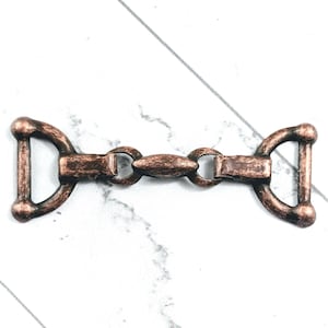 May include: A detailed, antique-style metal clasp in a copper finish. The clasp features a central oval piece connected to two D-shaped loops on either end. The design suggests a vintage or rustic aesthetic, suitable for various craft projects.