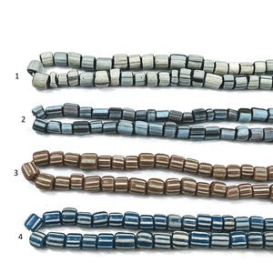 Puede incluir: Cuatro hebras de cuentas de vidrio, cada una con un patrón de color diferente. La hebra superior es blanca con rayas negras, la segunda hebra es azul con rayas negras, la tercera hebra es marrón con rayas blancas y la hebra inferior es azul con rayas blancas.