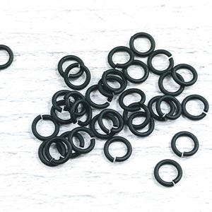 May include: A pile of black jump rings, a type of jewelry finding, on a white wooden surface.