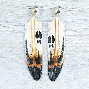 May include: Two white, brown and black painted feather pendants with silver bail loops. The feathers have a spotted pattern and black paw prints.