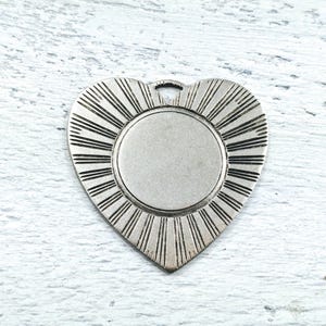 May include: Silver heart-shaped pendant with a circular, blank space in the center. The pendant has a sunburst design around the blank space.