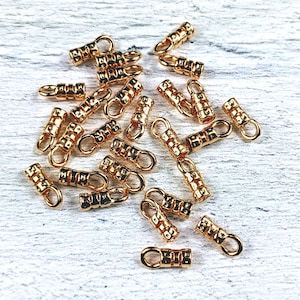 May include: A group of gold-toned metal crimp beads with a small loop on each end. The beads are scattered on a white wooden surface.