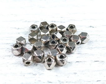 May include: A collection of silver-colored, faceted beads with a geometric design. Each bead has multiple sides and a hole running through the center. The beads are scattered on a white wooden surface.