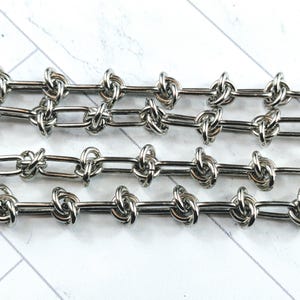 May include: Silver chain with a knotted design. The chain is made of metal links that are twisted together to create a knot-like appearance.