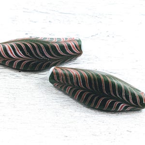 May include: Two green and pink glass beads with a white swirl pattern. The beads are oval shaped and have a glossy finish.