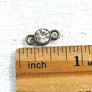 May include: A close-up of a small, antique gold-toned connector with a clear rhinestone in the center. The connector has two loops on either side of the rhinestone. The connector is measured against a ruler with the word "inch" printed on it.