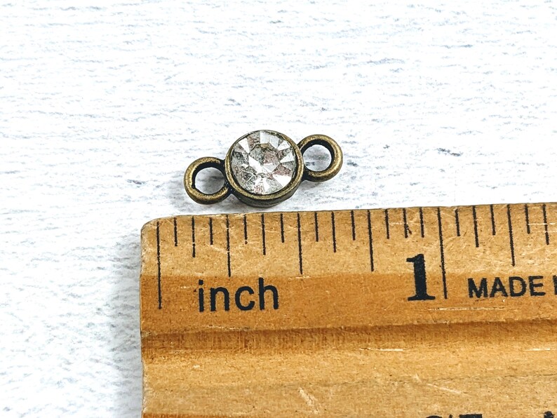 May include: A close-up of a small, antique gold-toned connector with a clear rhinestone in the center. The connector has two loops on either side of the rhinestone. The connector is measured against a ruler with the word "inch" printed on it.
