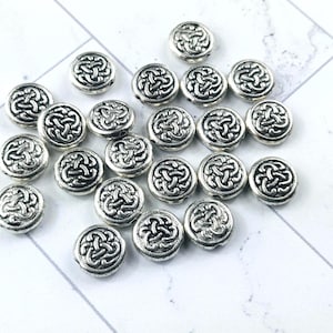 May include: A collection of silver-colored, round beads with a Celtic knot design. The beads are scattered on a white and gray surface. The beads are approximately 0.5 inches in diameter.