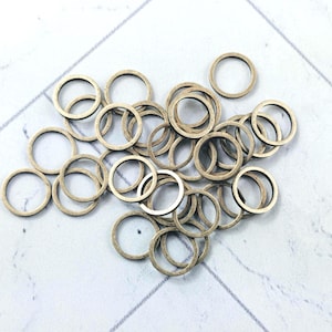May include: A collection of small, metallic-colored rings scattered on a white and gray surface. The rings appear to be made of metal and have a circular shape with a hole in the center. They are likely used for jewelry making or crafts.