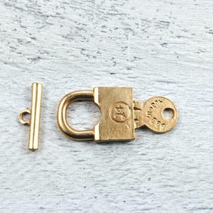 Puede incluir: Cierre de metal dorado con diseño de candado y una cerradura. El cierre tiene una pequeña barra separada que se desliza a través del candado para asegurarla.