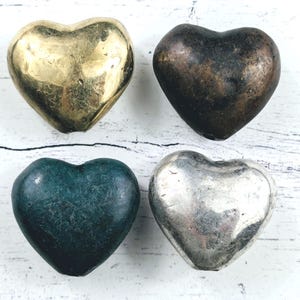 May include: Four heart-shaped objects in gold, bronze, teal, and silver. The hearts are smooth and metallic, with varying degrees of shine and patina. The objects are arranged on a white, weathered wooden surface.