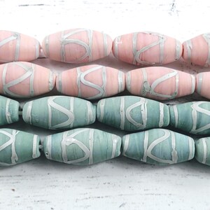 May include: Rows of oval-shaped beads in pink and teal. Each bead has a silver-colored, abstract design. The beads are strung together, suggesting they are for jewelry making or crafting. The beads are displayed on a white surface.