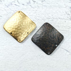 Puede incluir: Dos abalorios cuadrados de metal con textura martillada. Un abalorio es dorado y el otro es negro.