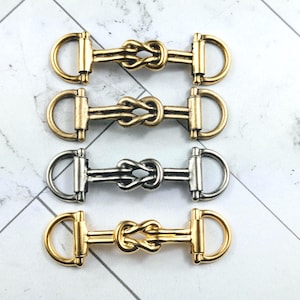 Peut inclure: Quatre connecteurs de mors de bride en métal, dans des tons or et argent. Chaque connecteur présente un motif de nœud au centre et des anneaux en D à chaque extrémité. Les mors sont disposés sur une surface à motifs de marbre blanc et gris.