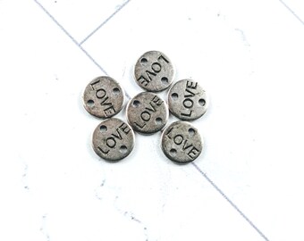 May include: Six small, round, silver-colored charms with the word "LOVE" stamped on each. Each charm has three small holes. The charms are arranged on a white surface with a subtle pattern.