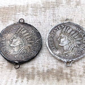 May include: Two silver-toned pendants featuring an image of a Native American woman with a feathered headdress. The pendants are both inscribed with "United States of America".