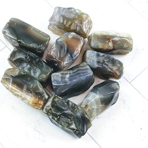 May include: A collection of tumbled stones in shades of gray, brown, and cream. The stones have a smooth, polished appearance and varying shapes. The natural stones are arranged on a white surface, showcasing their unique patterns and textures.