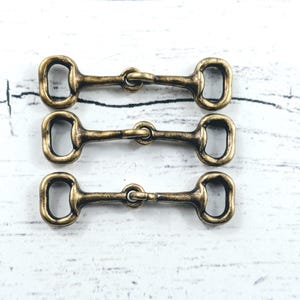 Puede incluir: Tres conectores de bocado de caballo de metal de color latón antiguo. Cada bocado tiene una pieza central rectangular con un anillo en cada extremo y un anillo de conexión en el medio. Los bocados están dispuestos horizontalmente.