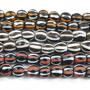 Könnte beinhalten: Eine Sammlung von gestreiften Glasperlen in Schwarz, Weiß und Orange. Die Perlen sind in drei Reihen angeordnet, wobei die obere Reihe orange-weiße Streifen, die mittlere Reihe schwarz-weiße Streifen und die untere Reihe schwarz-weiß-orange Streifen aufweist.