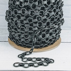 May include: A spool of black chain with a section of chain lying on a white wooden surface. The chain is made of metal and has a matte finish.