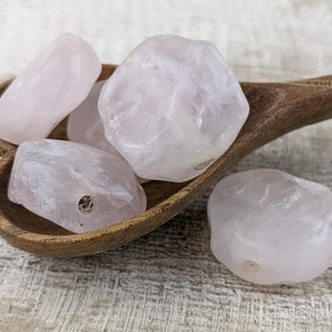 May include: Four pale pink rose quartz stones with holes drilled through them, resting in a wooden bowl. The stones are smooth and have a natural, irregular shape.