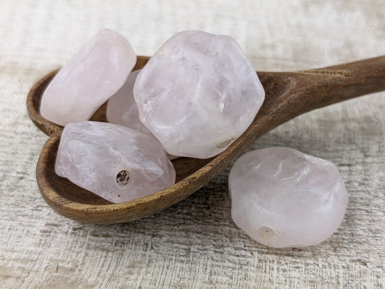 May include: Four pale pink rose quartz stones with holes drilled through them, resting in a wooden bowl. The stones are smooth and have a natural, irregular shape.