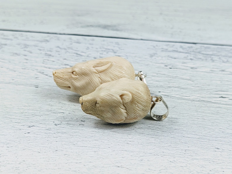 May include: Two ivory-colored bear head pendants with silver-colored loops. The pendants are carved with detailed features, including the bear's snout, eyes, and fur. The pendants are displayed on a light-colored wooden surface.