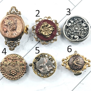 May include: A selection of six antique buttons in diverse shapes and designs. The buttons showcase intricate details, including floral patterns, a rabbit, and a classical figure. The buttons are made of metal and have a vintage aesthetic.