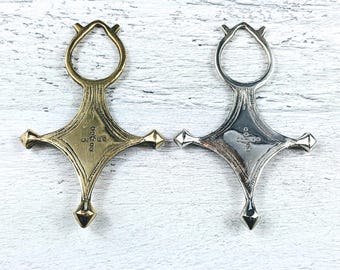 May include: Two silver and gold pendants in the shape of a cross with a loop at the top. The cross is a traditional Ethiopian design with a central diamond shape and four points. The pendants are on a white wooden background.