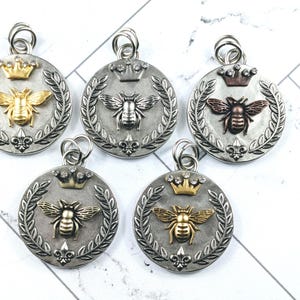 May include: Five silver-toned pendants with bee and crown designs. Each pendant features a bee in gold, silver, or bronze tones, surrounded by a laurel wreath and topped with a crown. The pendants are round with a loop for a chain.