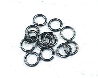 May include: A selection of dark grey metal jump rings, some open and some closed, on a white surface. The circular rings have a metallic sheen, ideal for jewellery making or crafts.