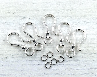 May include: Six silver-toned lobster claw clasps with a round bead at the top. The clasps are arranged in a semi-circle on a white wooden surface. There are also six silver-toned jump rings scattered around the clasps.