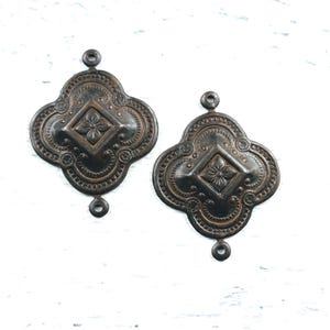 May include: Two antique-looking metal embellishments with a four-leaf clover shape and a raised diamond-shaped design in the centre. Each embellishment has three holes for attaching.