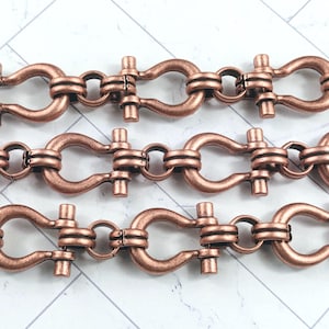 May include: Close-up of three rows of copper-colored chain links. Each link has a U-shaped design connected by a series of rectangular bars. The links are arranged in a repeating pattern, creating a textured and industrial aesthetic.
