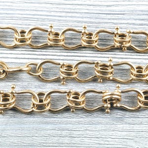 May include: A gold-toned metal chain necklace featuring a series of interlocking U-shaped links connected by small circular rings. The chain has a matte finish and is arranged in three horizontal rows, showcasing a repeating pattern of metal loops.