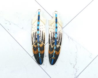 May include: A pair of white feather earrings with blue, black, and orange painted details. The feathers are long and narrow, with a loop at the top for hanging.