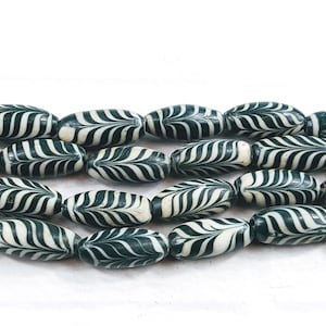 Puede incluir: Primer plano de cuentas verdes oscuras y blancas. Cada cuenta es ovalada, con una base verde oscuro y un diseño blanco en forma de hoja. Las cuentas están ensartadas, perfectas para crear joyas.