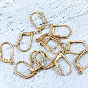May include: A collection of gold-colored earring lever back findings, scattered on a light-colored, textured surface. The findings are in various orientations, showcasing their curved and hinged design, ideal for jewelry making.