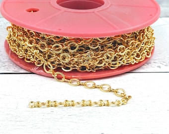 Puede incluir: Un carrete de cadena de color dorado, enrollado alrededor de un carrete de plástico rosa. La cadena es de estilo delicado, de eslabones ovalados.