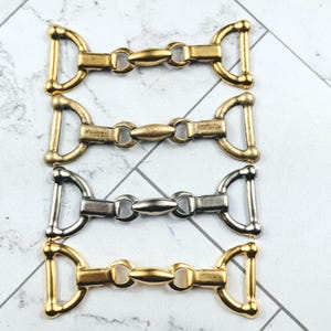 May include: Four metal bit connectors in gold, antique brass, and silver finishes. Each connector has a D-ring on each end and a central oval link. The connectors are arranged in a vertical stack.