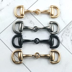 May include: Four metal horse bit charms in gold, silver, and black. The charms are arranged horizontally, showcasing their design and colour variations. The bits have a classic equestrian style, with a central ring connecting the two D-shaped ends.