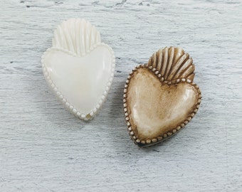 May include: Two heart-shaped decorative objects. One is off-white, the other is brown. Both have a textured, fan-like design at the top and a beaded border. The hearts are on a light-colored, textured surface.