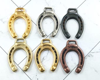 May include: Six metal horseshoe charms in gold, silver, black, and bronze finishes. Each horseshoe has a loop for attachment. The charms are displayed on a white surface with a subtle geometric pattern. Ideal for jewellery making or crafts.