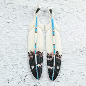 May include: Two feather pendants with silver-colored bails. The feathers are primarily cream-colored with black, pink, and brown accents. A vertical line of turquoise and brown beads runs down the center of each feather.