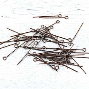 May include: A collection of copper-colored eye pins scattered on a white, textured surface. Each pin has a straight wire body with a looped eye at one end. The pins are of varying lengths and are suitable for jewelry making and crafting.