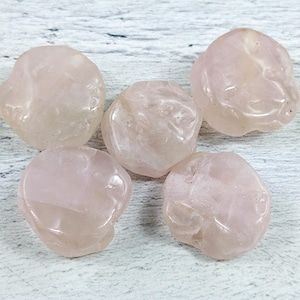 Könnte beinhalten: Fünf glatte, blassrosa Rosenquarzsteine, jeder mit einer einzigartigen Form und Textur, sind auf einer weißen Holzoberfläche angeordnet.