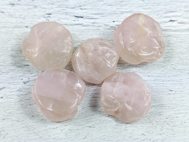 May include: Five smooth, pale pink rose quartz stones, each with a unique shape and texture, are arranged on a white wooden surface.