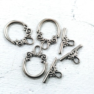 May include: Silver-toned metal clasps with ornate designs. The clasps are in the shape of a circle and a T.